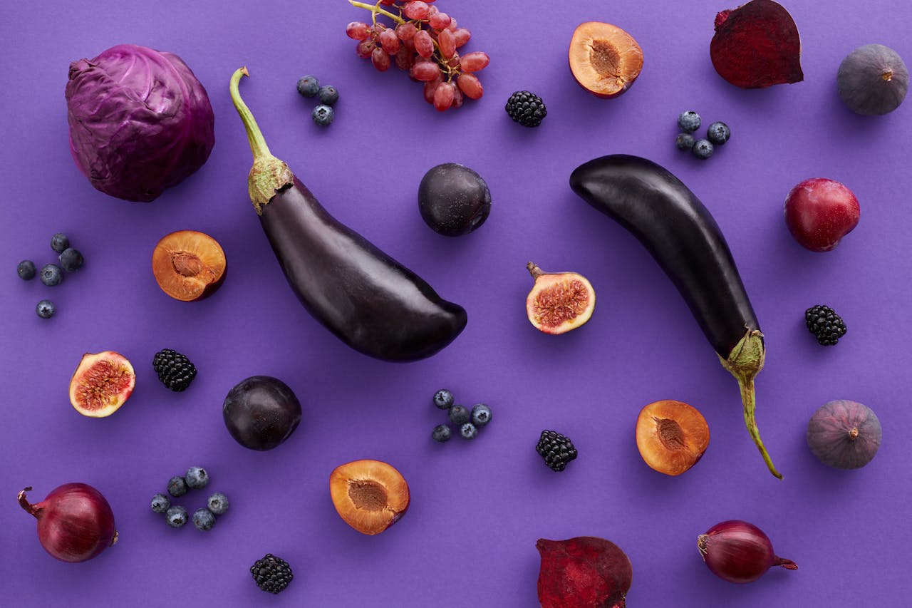 Vibrant top view of fresh fruits and vegetables like eggplants, figs, and plums laid on a purple background.